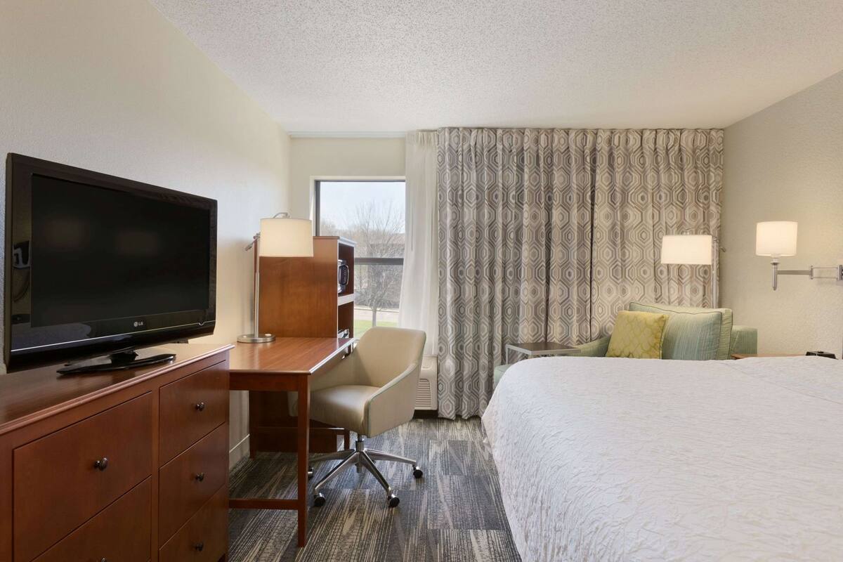 Ein Hotelzimmer mit Flachbildfernseher, Schreibtisch mit Lampe, Stuhl, Bett mit Kissen und Fenster mit gemusterten Vorhängen.