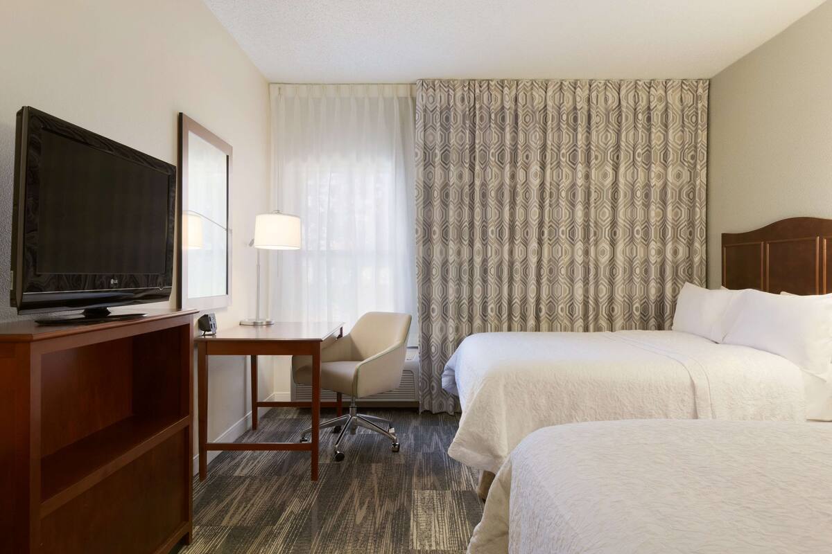 Ein Hotelzimmer mit zwei Betten, einem Fernseher auf einem Holzständer, einem Schreibtisch mit Stuhl und gemusterten Vorhängen.