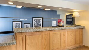 A reception area with a wooden counter, framed pictures, a welcome sign, a lamp, and a vending machine. - Hampton Inn Zanesville (Zanesville)