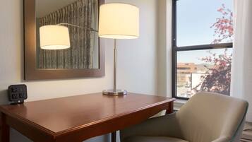 Hotelkamer met een houten bureau, een lamp, een stoel, een spiegel en een raam met uitzicht op gebouwen en bomen.