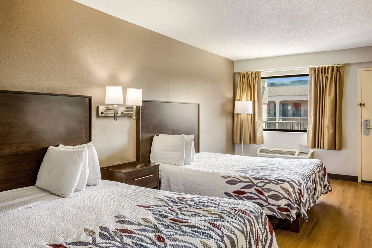 Habitación de hotel con dos camas, una mesita de noche, una lámpara, una ventana con cortinas y vistas a un edificio.