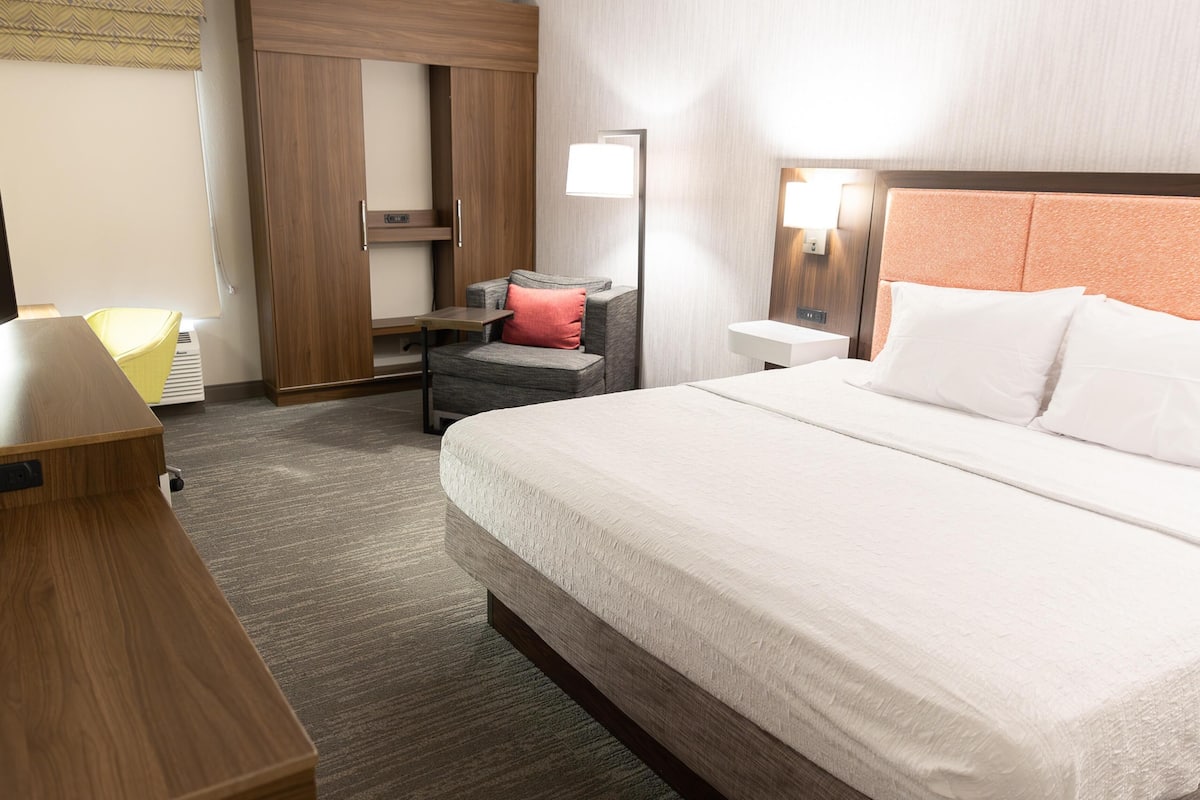 A hotel room with a bed, a desk, a chair, a wardrobe, and a nightstand with a lamp.