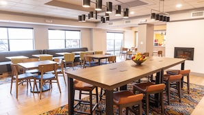 A modern dining area with multiple tables and chairs, a central fruit bowl, and a fireplace. - Hampton Inn Williamsport-Downtown (Williamsport)