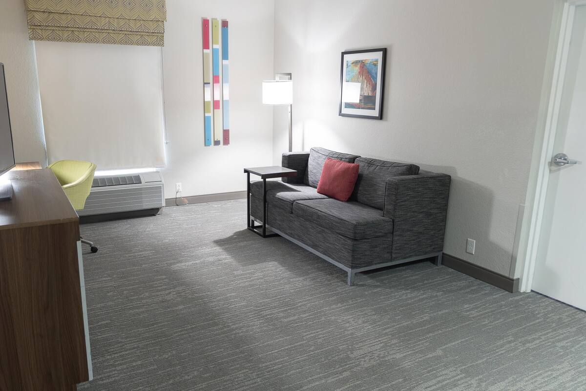 A room with a grey sofa, a small table, a standing lamp, a yellow chair, and a framed picture on the wall.