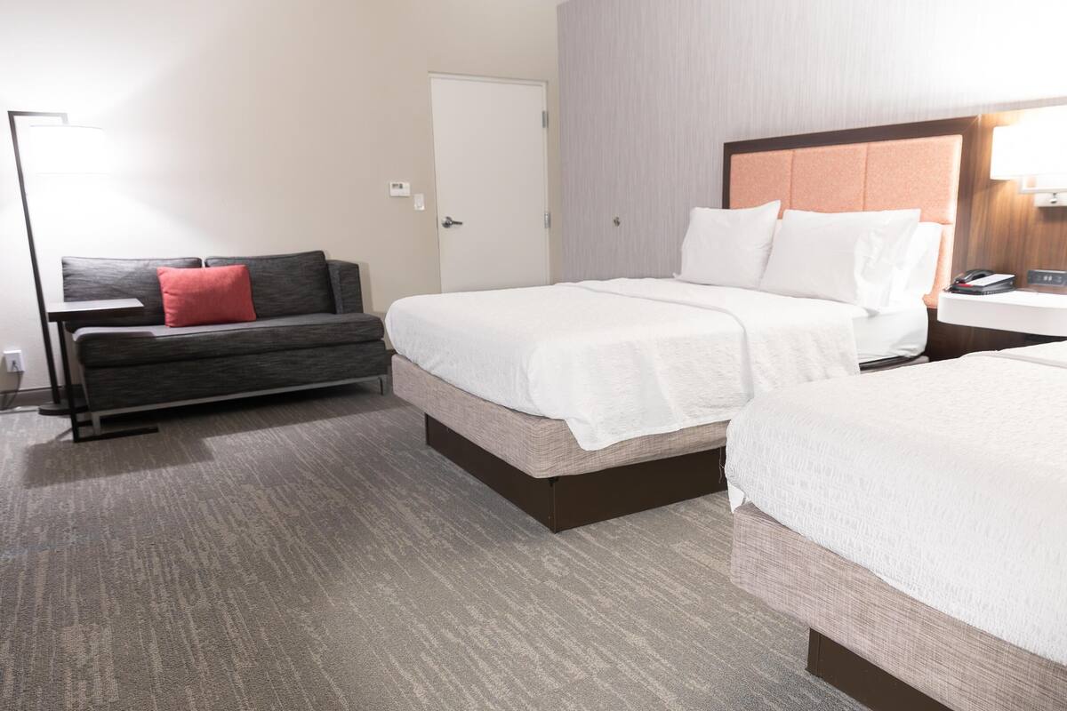 A hotel room with two beds, a sofa, a bedside table, and a lamp.