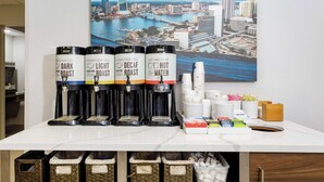 Una estación de café con cuatro dispensadores de café: tostado oscuro, tostado claro, descafeinado y agua caliente. Hay pilas de vasos desechables y una variedad de cápsulas de café. Se ve un cuadro de un paisaje urbano en la pared detrás de la estación.
