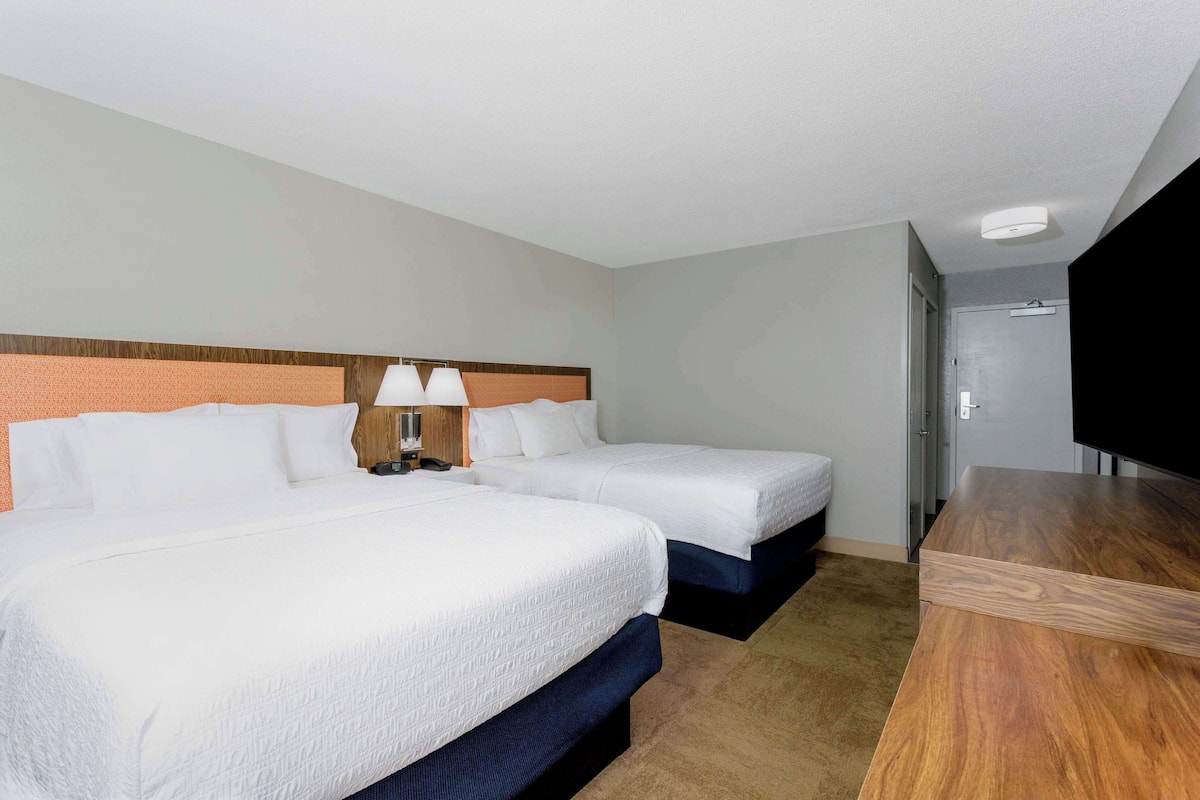 A hotel room with two beds, a wooden headboard, a desk with a chair, and a flat-screen TV.