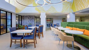 Un comedor interior moderno con mesas y sillas, un sofá verde con cojines naranjas, y una pared con diseño de hojas.