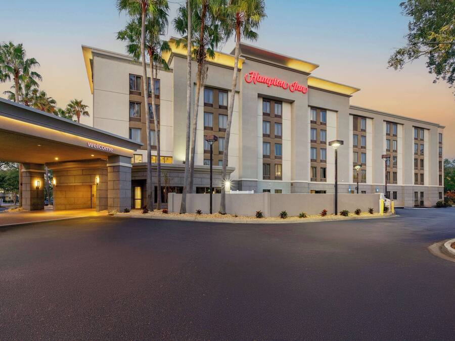 A modern hotel with a prominent "Hampton Inn" sign, a well-lit entrance, and a landscaped area with palm trees.