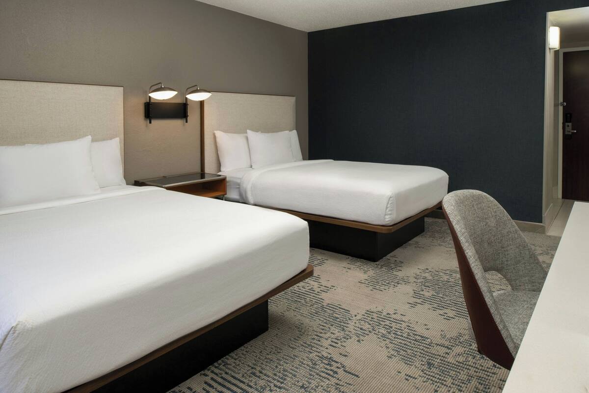A hotel room with two beds, a desk, and a chair. There are two bedside tables with lamps.