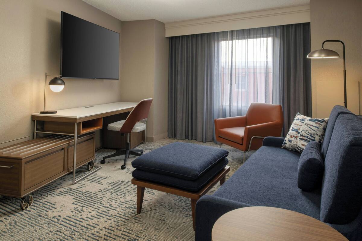 A hotel room with a desk, a chair, a sofa, a TV mounted on the wall, and a window with curtains.