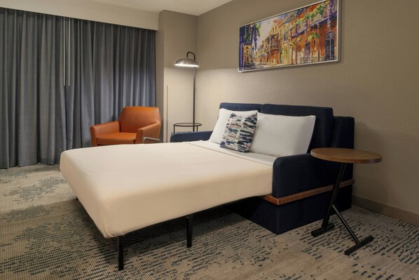 A hotel room with a large bed, a chair, a small table, and a painting on the wall.