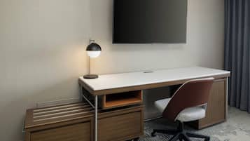 Quarto de hotel com uma escrivaninha branca, uma luminária moderna, uma TV de tela plana, uma cadeira marrom e um pequeno armário de madeira.