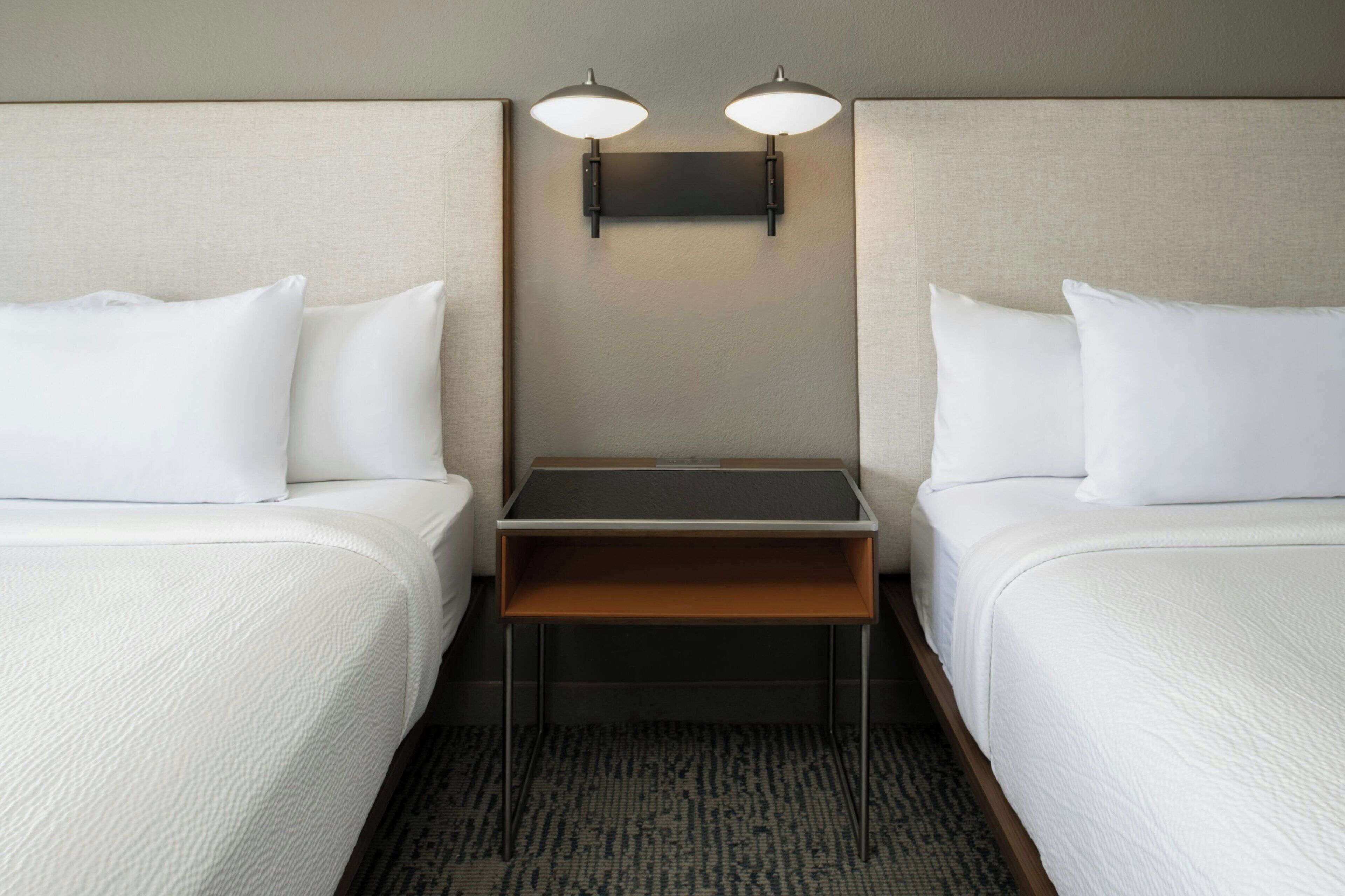 A hotel room with two beds, a small table, and a wall-mounted light fixture.