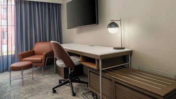Una habitación de hotel con escritorio, una silla, televisor montado en la pared y lámpara.