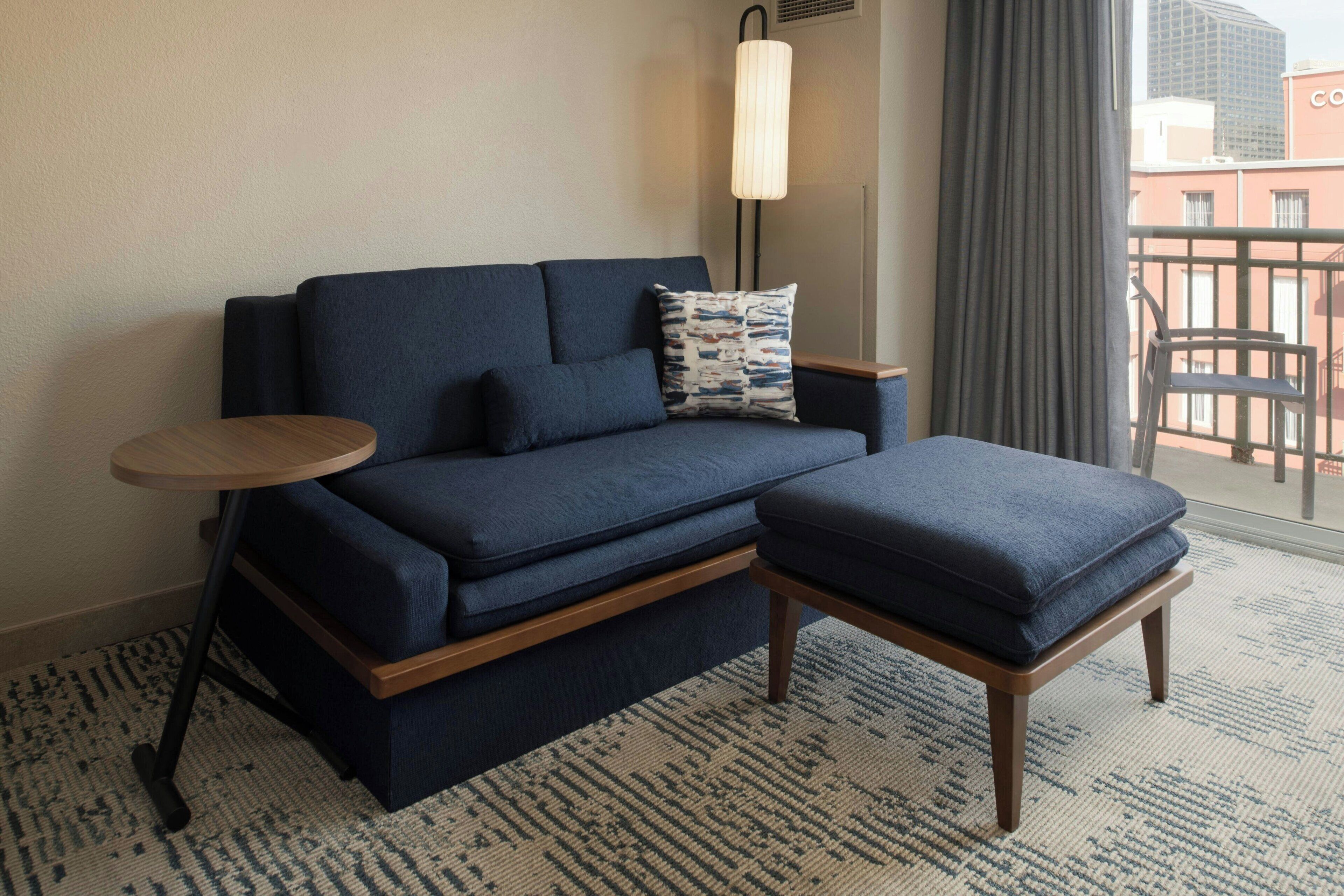 Et moderne hotelværelse med en mørkeblå sofa, tilhørende skammel, et lille træsidebord og en gulvlampe.