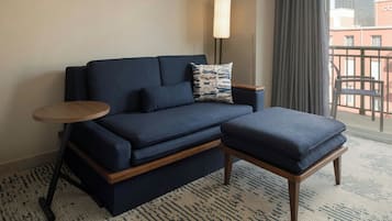Une chambre d’hôtel moderne avec un canapé bleu foncé, un pouf assorti, une petite table d’appoint en bois et une lampe sur pied.
