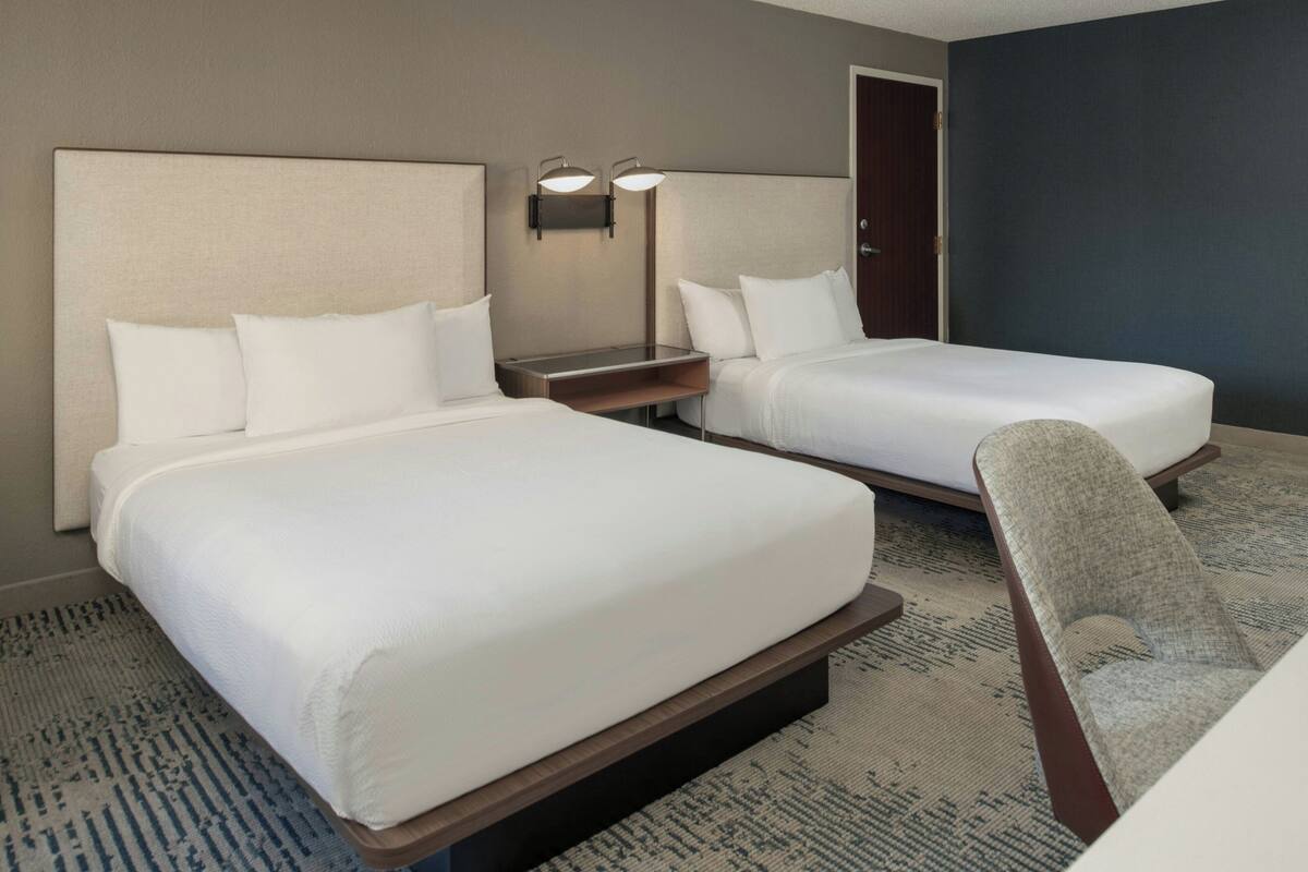 A hotel room with two beds, a desk, a chair, and a nightstand with lamps.