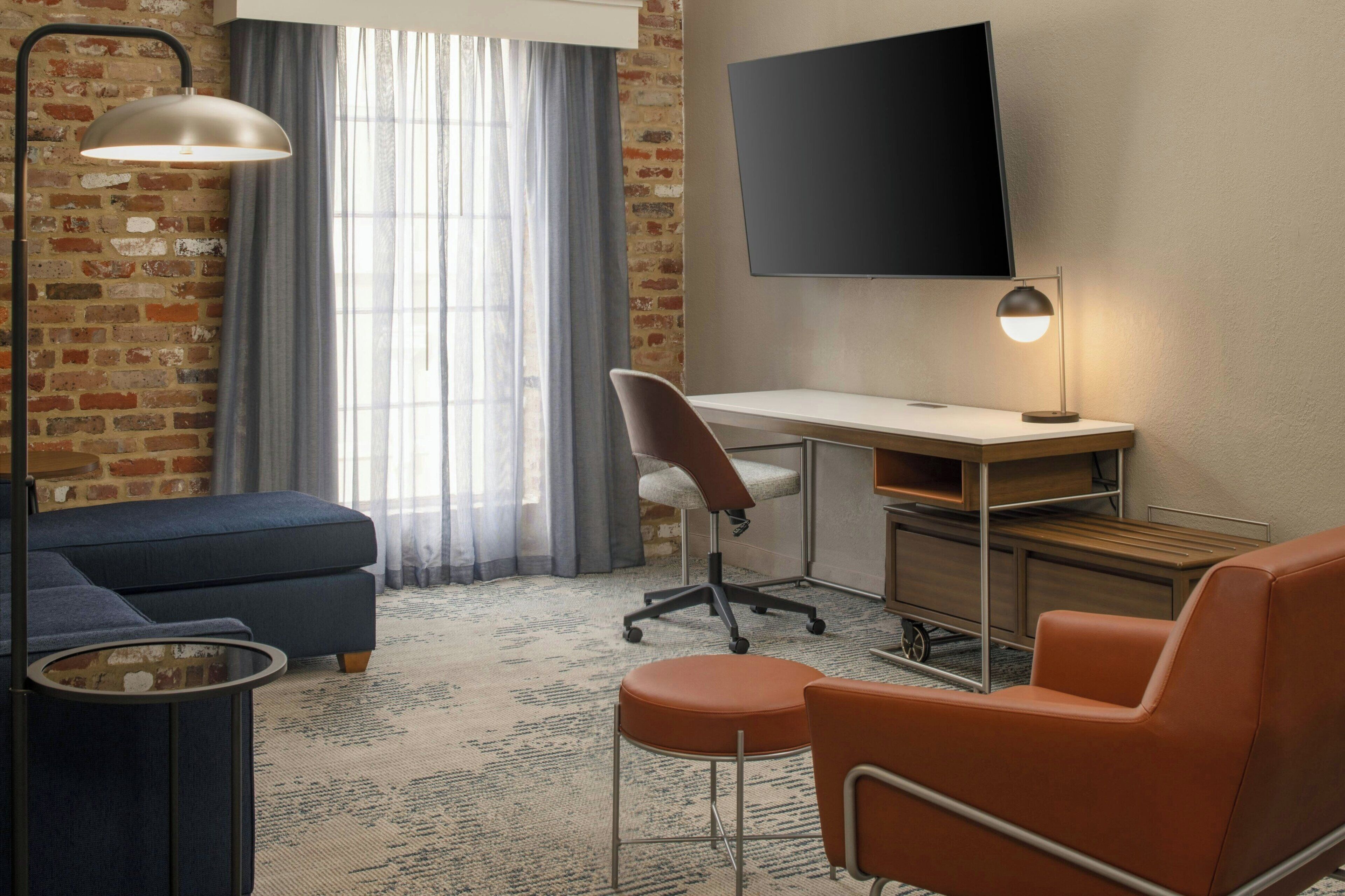 Una habitación de hotel moderna con pared de ladrillo, un televisor de pantalla plana grande montado en la pared, un escritorio con una silla, un sofá y un sillón naranja.