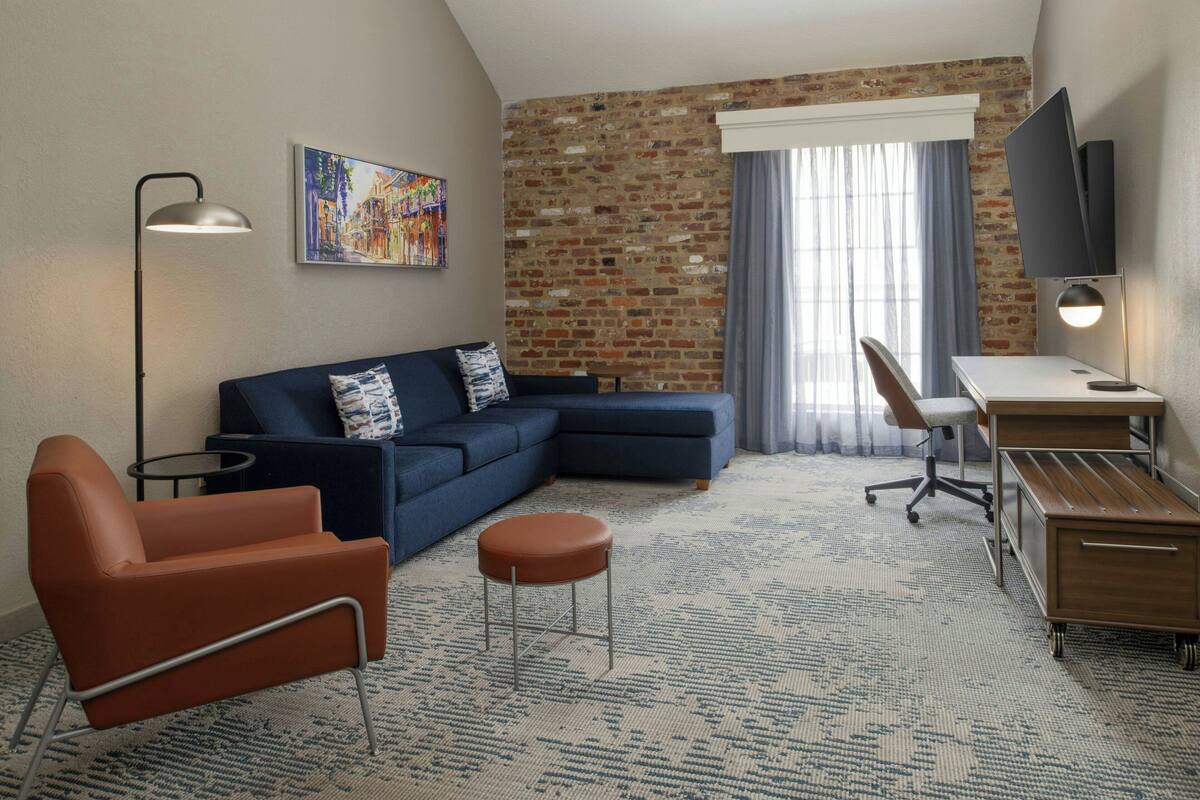 A modern hotel room with a sectional sofa, a single armchair, a small round ottoman, a desk with a chair, a flat-screen TV, and a large window with curtains.