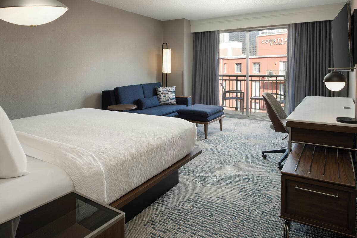 A hotel room with a large bed, a desk, a chair, a sofa, a balcony with a view of a building, and a large window.