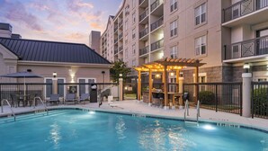 Una piscina con una pérgola y sillas de descanso, rodeada por una valla y un edificio de varios pisos con balcones.
