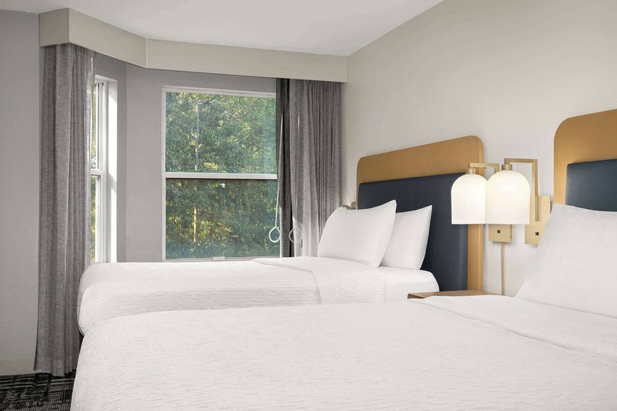 A hotel room with two beds, a large window with curtains, and a nightstand with a lamp.