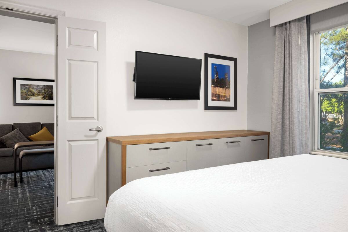 A hotel room with a bed, a TV mounted on the wall, a desk with drawers, a sofa, and a framed picture.