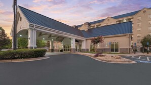 Entrada de hotel con galería techada, zona de estacionamiento y jardín ajardinado.