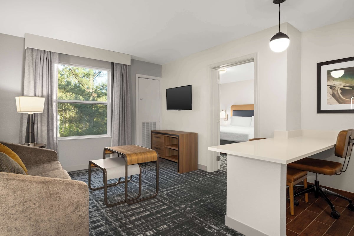 A compact hotel room with a sofa, a small desk, a chair, a TV, and a large window with curtains.