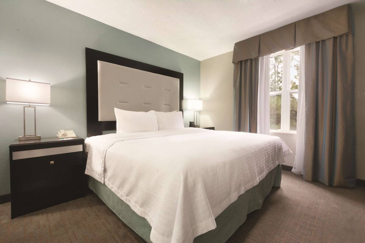 A hotel room with a large bed, two bedside tables, a nightstand, a window with curtains, and a view of trees.