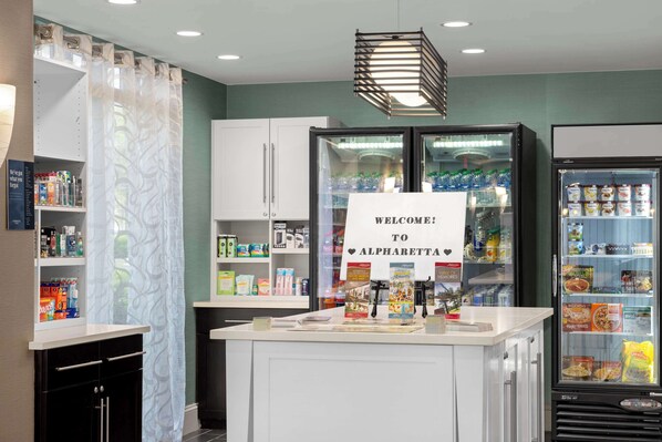 A kitchen area with a welcome sign, a refrigerator stocked with beverages, and shelves with various food items. - Homewood Suites by Hilton Atlanta-Alpharetta (Alpharetta)