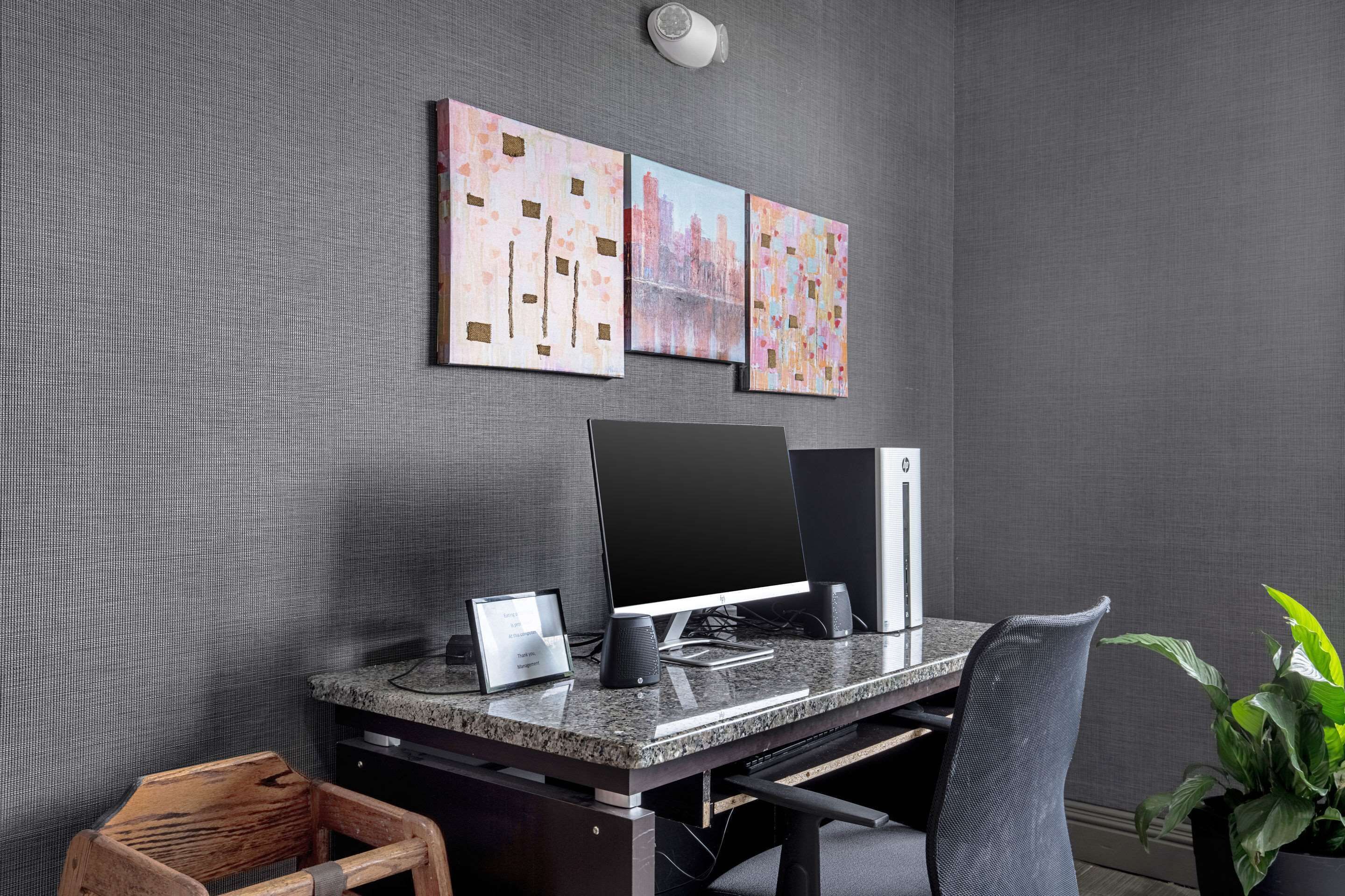 A modern office space with a desktop computer, two monitors, a keyboard, and a mouse on a granite desk. There is a wooden chair and a potted plant in the corner.