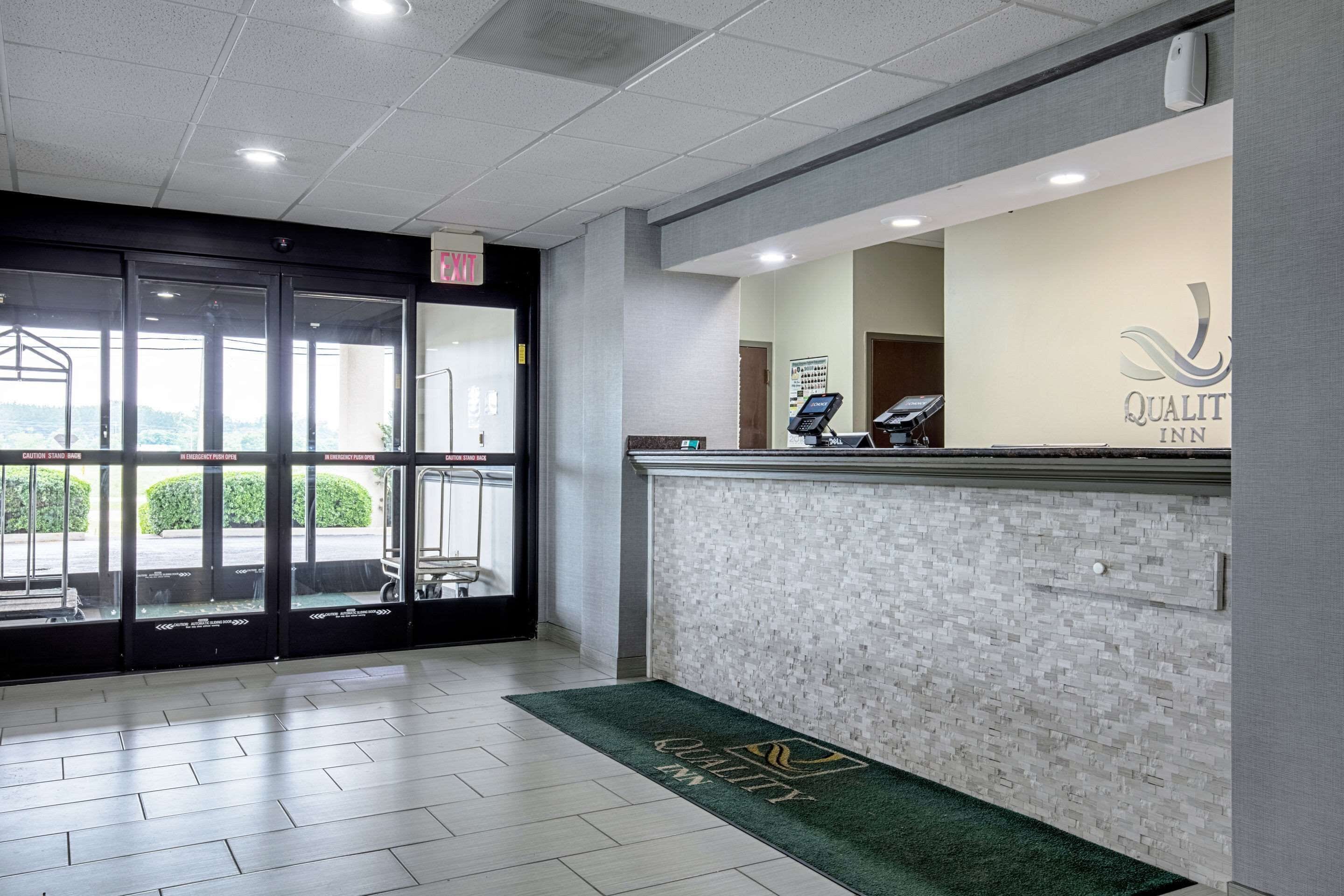 Un hall d’accueil d’hôtel avec une entrée dotée d’une porte en verre, un comptoir de réception et un tapis vert orné du logo de l’hôtel.
