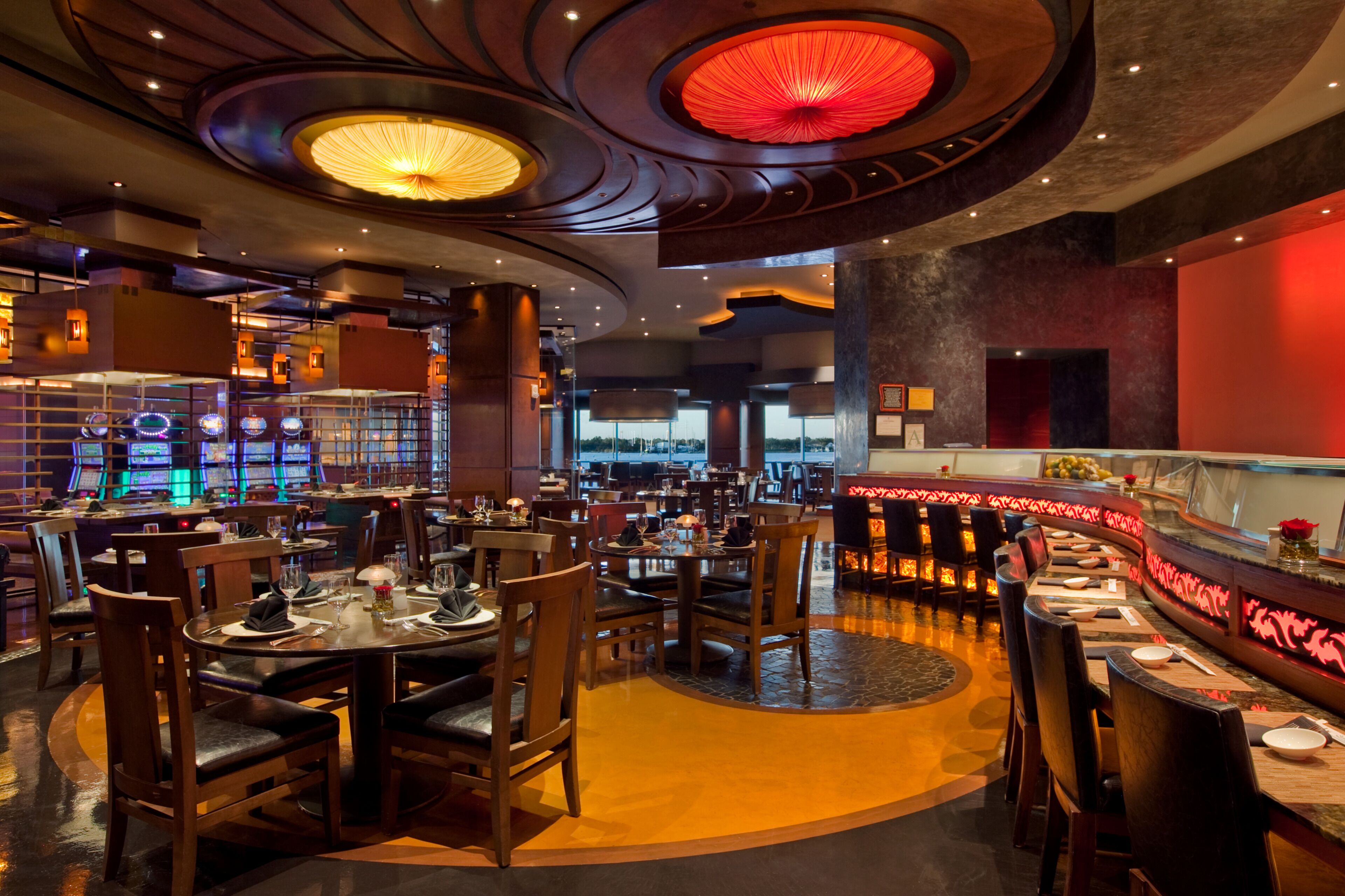 A restaurant with round tables and booths, a curved bar, and a view of the cityscape through large windows.