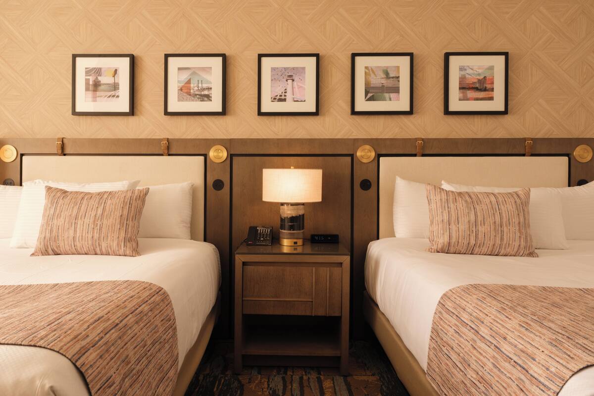 Habitación de hotel con dos camas, una mesita de noche con lámpara y cuadros en la pared.