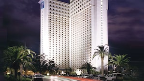 Un hotel de gran altura con un llamativo letrero 'IP', rodeado de palmeras y una calle concurrida con estelas de luz.