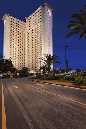 A high-rise hotel with a prominent vertical sign on the facade, a well-maintained road in front, and palm trees lining the roadside.