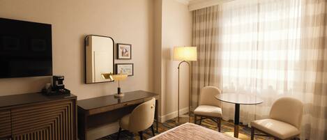 A hotel room with a bed, a desk with a chair, a small table with two chairs, a TV, a lamp, and a large window with curtains.