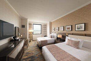 A hotel room with two beds, a desk, a TV, and a view of the ocean.