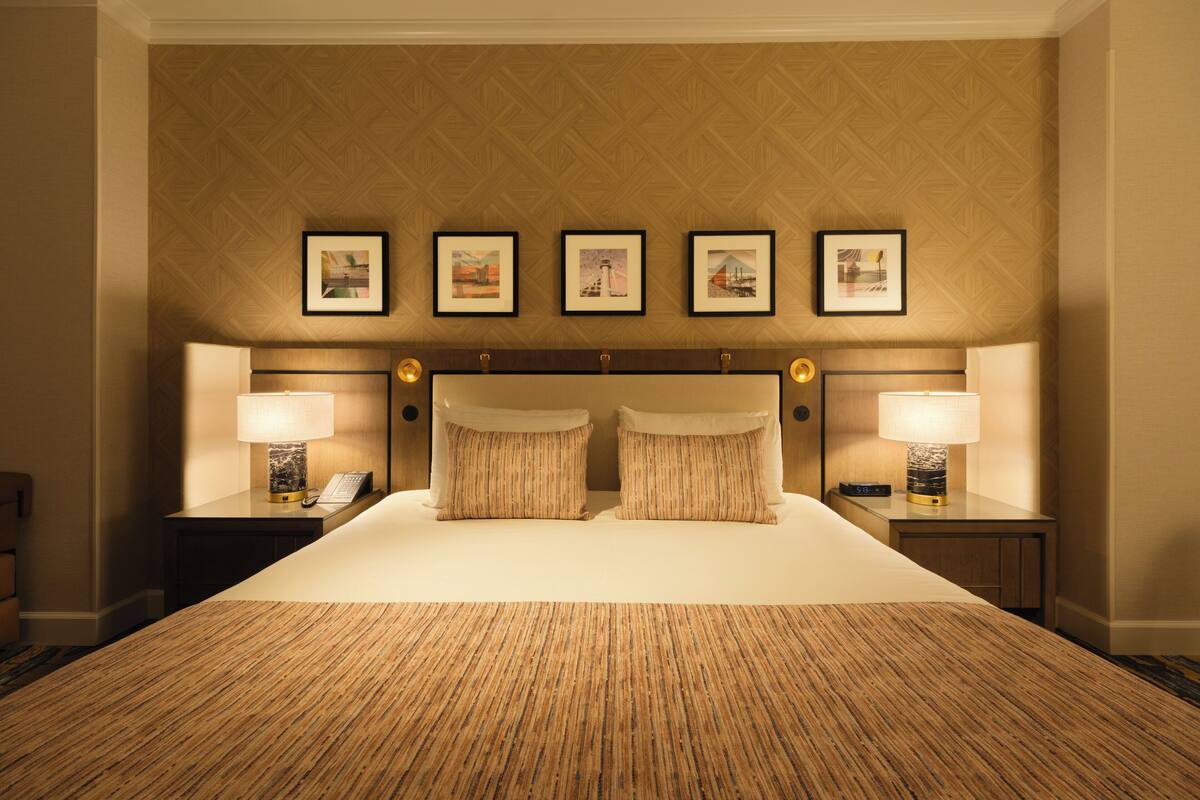 Una habitación de hotel con una cama grande, dos mesitas de noche con lámparas y un cabecero con cuadros enmarcados.