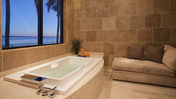 Un baño con bañera, ventana con vistas al mar y pared de azulejos color beige.