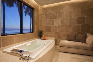 Un baño con bañera, ventana con vistas al mar y pared de azulejos color beige.