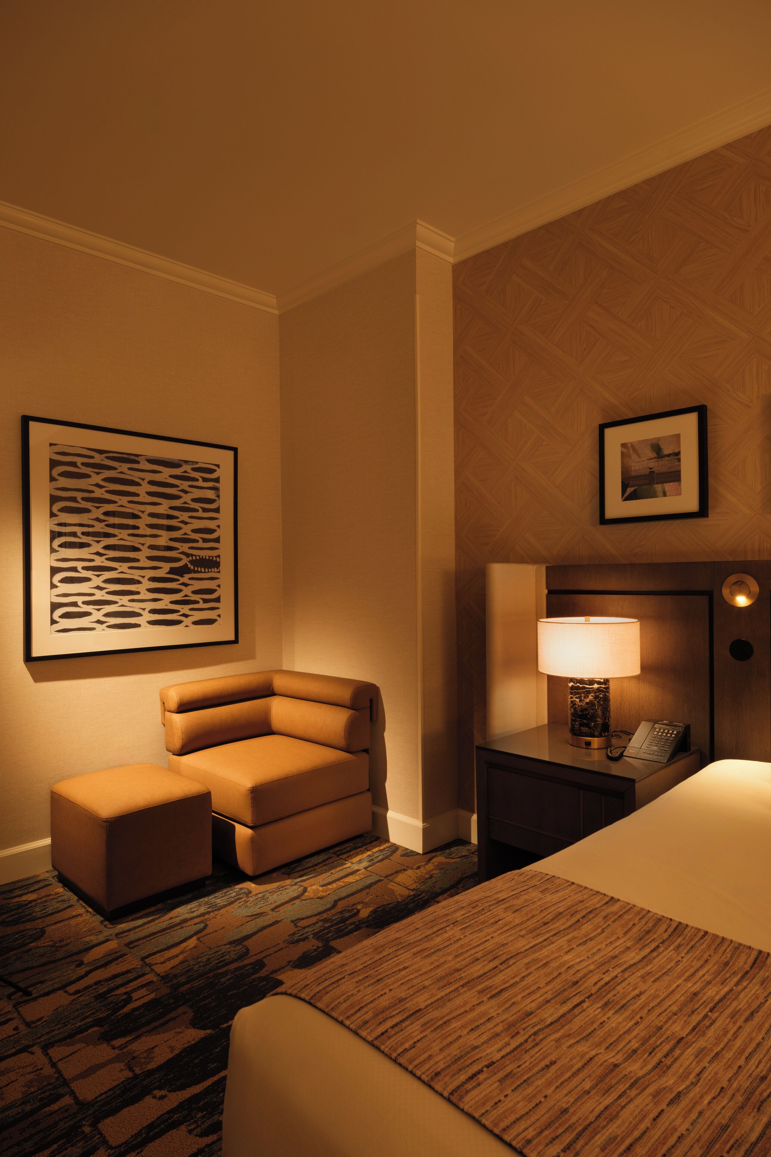 Een hotelkamer met een bed, een nachtkastje met lamp, een stoel en een kunstwerk in een lijst aan de muur.