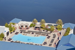 An aerial view of a resort with a large swimming pool surrounded by sun loungers and umbrellas, palm trees, and a clear view of the water.