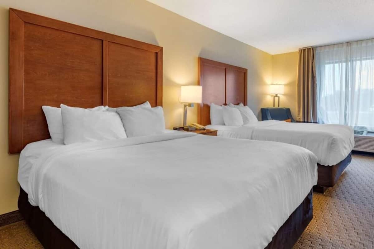 A hotel room with two beds, wooden headboards, bedside lamps, a small table, and a view of the outdoors.