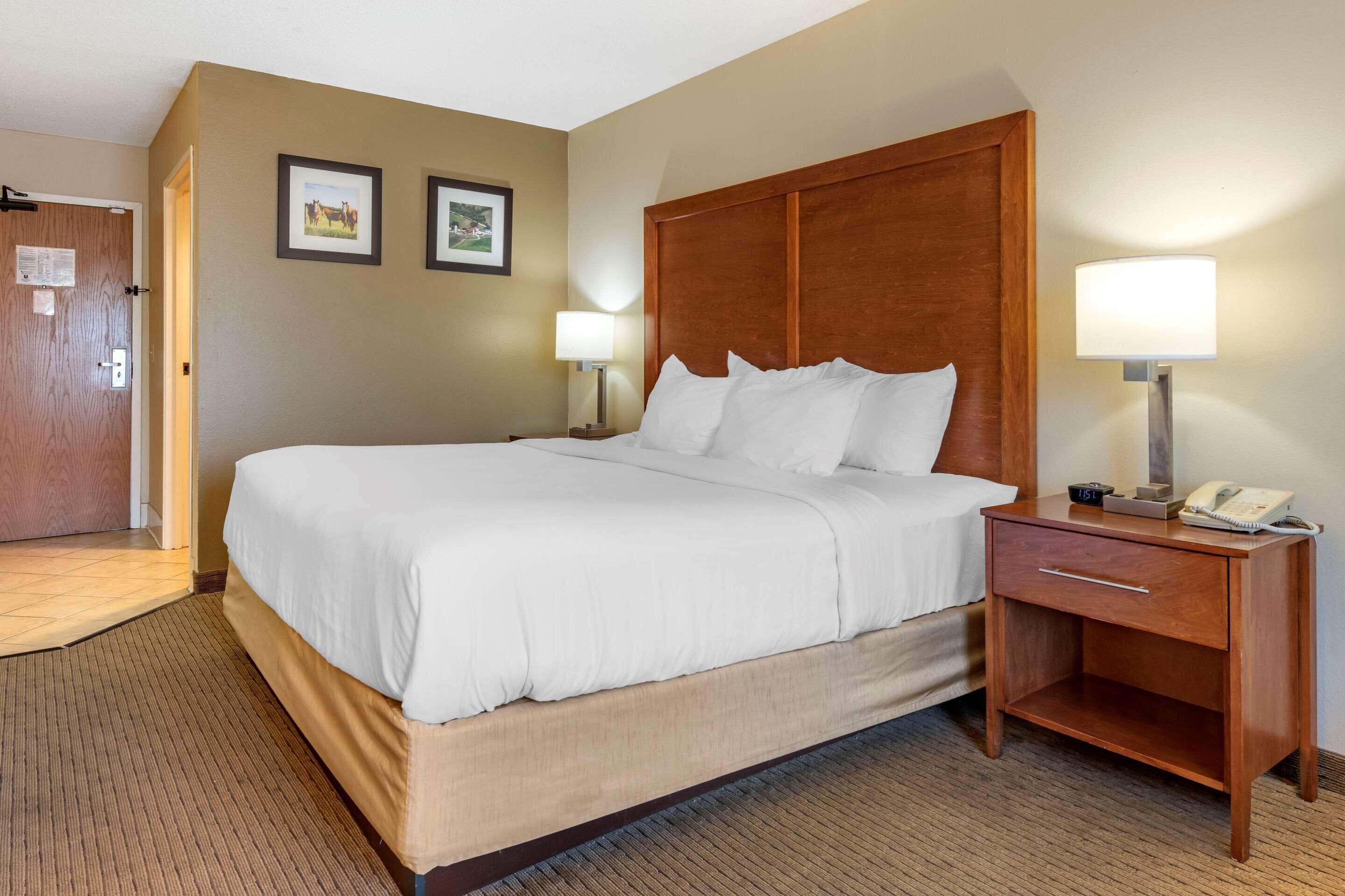 Une chambre d’hôtel avec un grand lit, deux tables de chevet avec des lampes, une petite table en bois et une porte avec une pancarte.