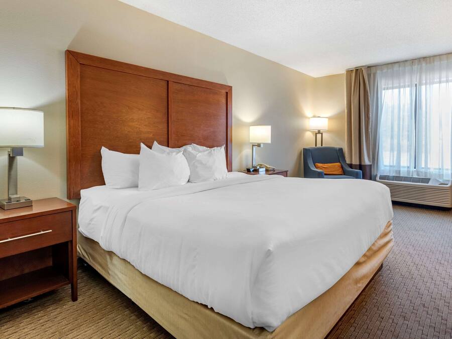 Una habitación de hotel con una cama grande, cabecero de madera, mesitas de noche con lámparas, una silla y un ventanal con cortinas.