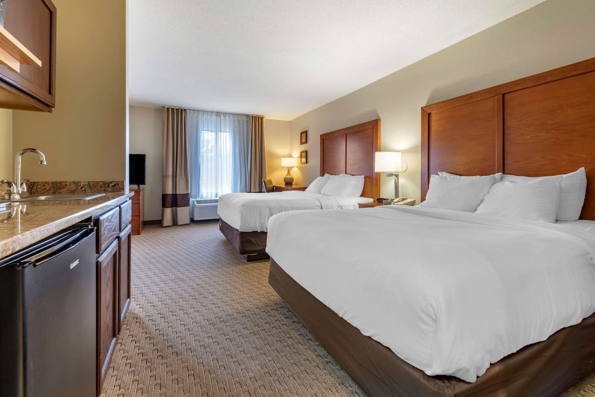 A hotel room with two beds, a kitchenette, a bathroom, and a large window with curtains.