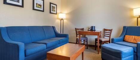 A hotel room with a blue sofa, a wooden coffee table, a small ottoman, a dining table with chairs, and two wall-mounted lamps.